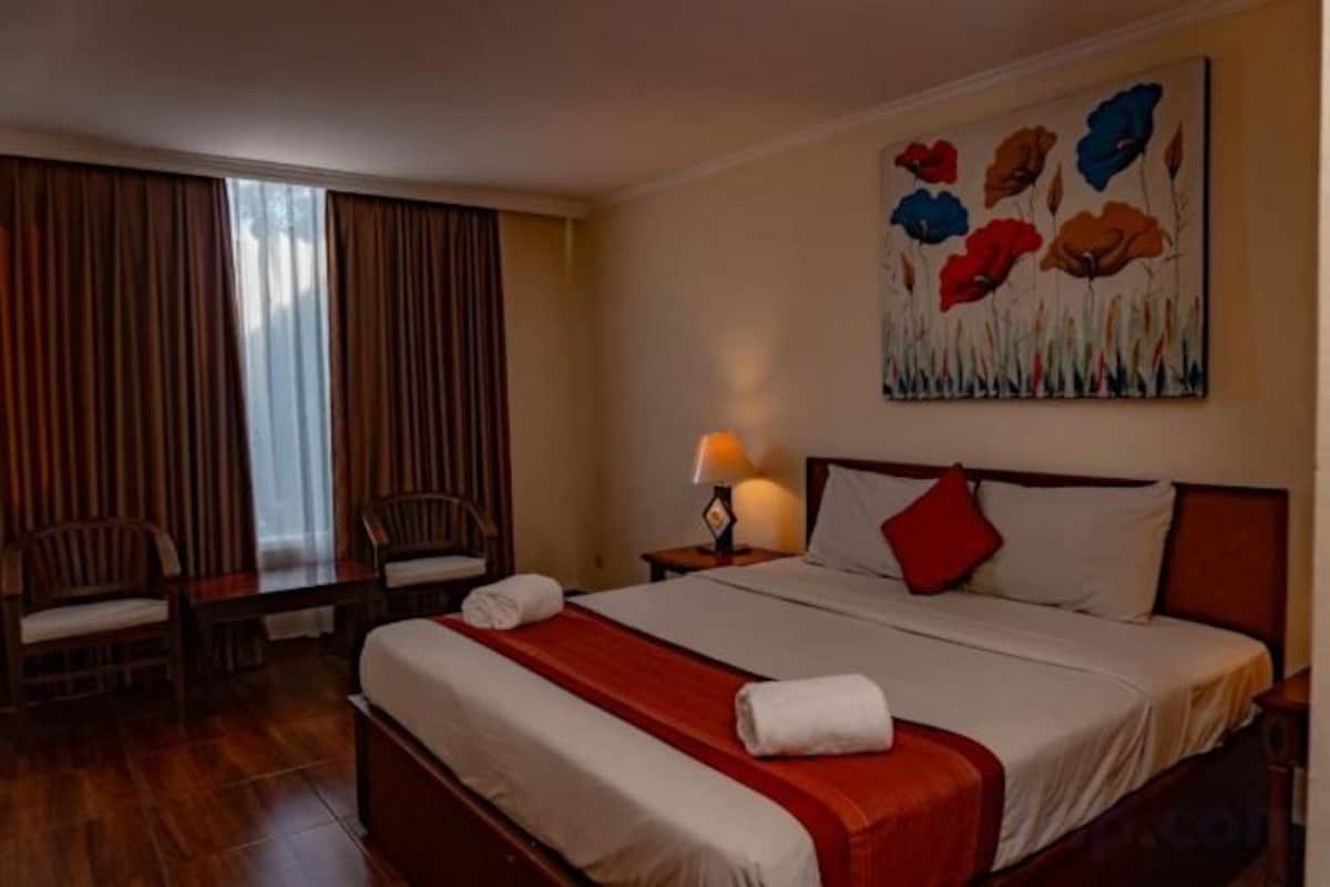 Habitación de hotel con una cama, dos sillas, una mesita de noche con lámpara y un cuadro en la pared.