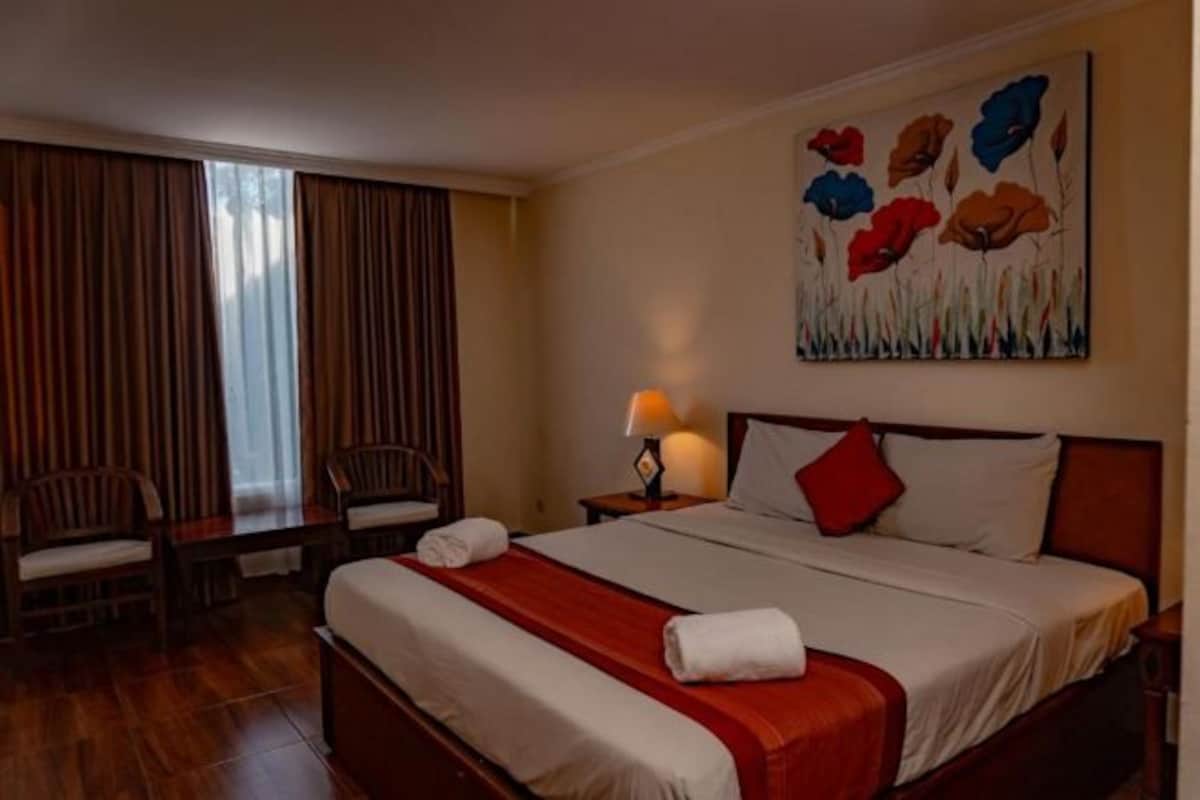 A hotel room with a bed, a desk, two chairs, a lamp, and a painting on the wall.