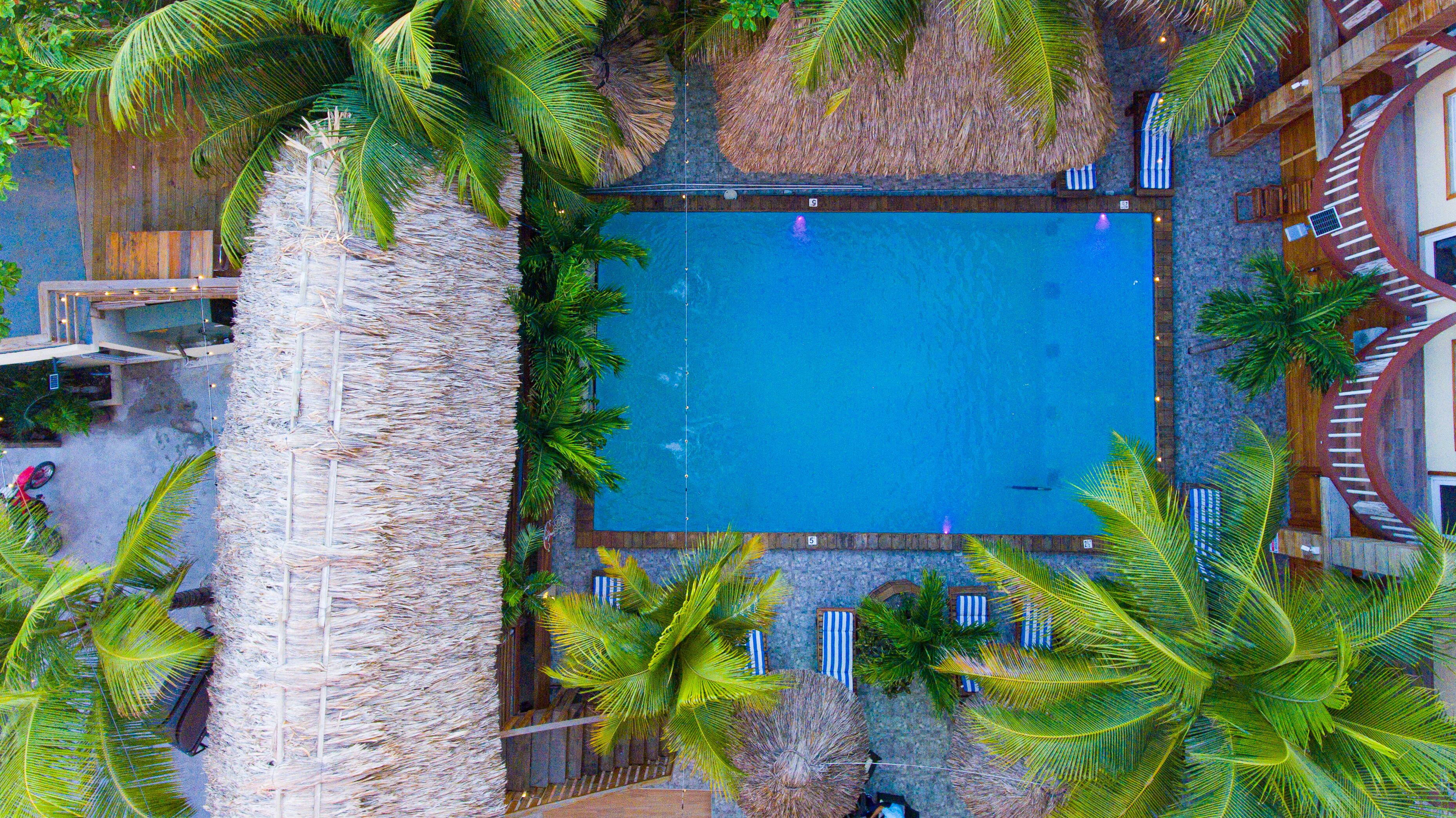 An aerial view of a resort with a central swimming pool surrounded by palm trees and a wooden deck.