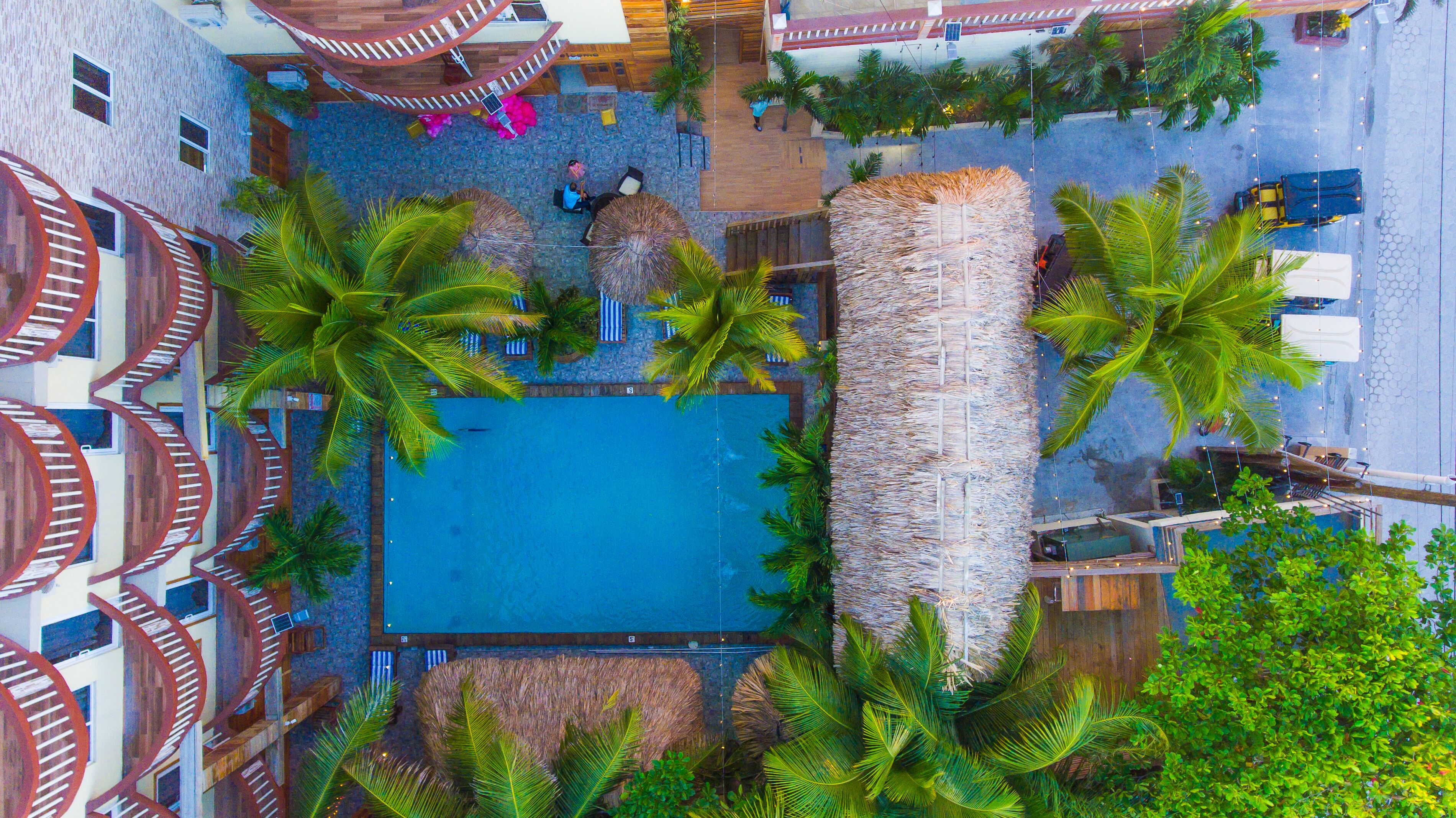 Vista aérea de un resort con una piscina central, rodeado de palmeras y sombrillas de paja.