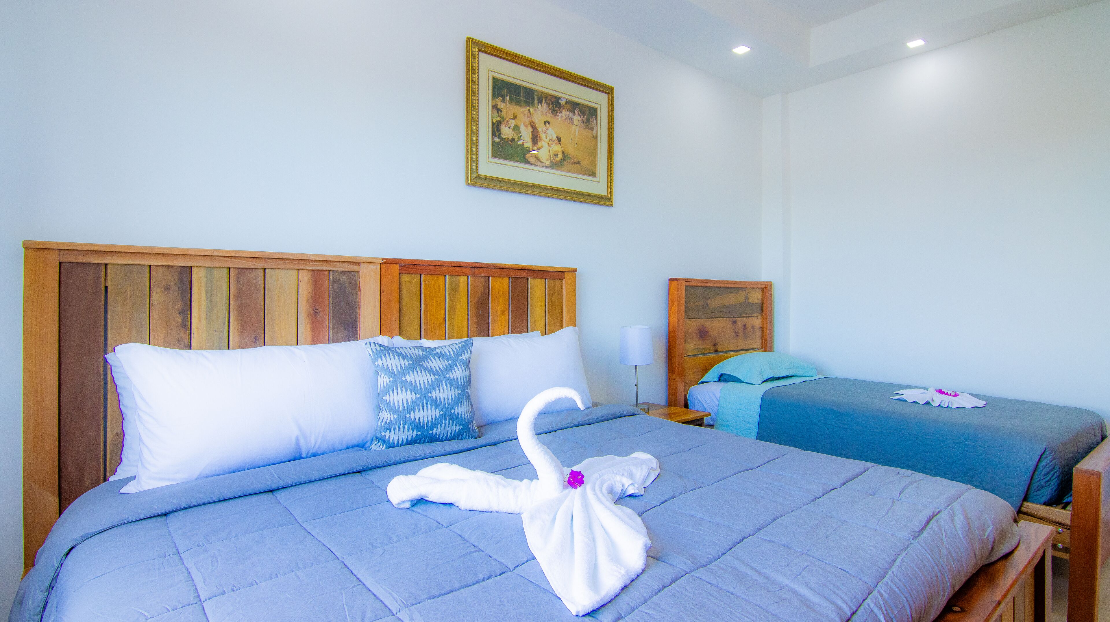 A hotel room with a large bed, a smaller bed, wooden headboards, a painting on the wall, and a nightstand with a lamp.