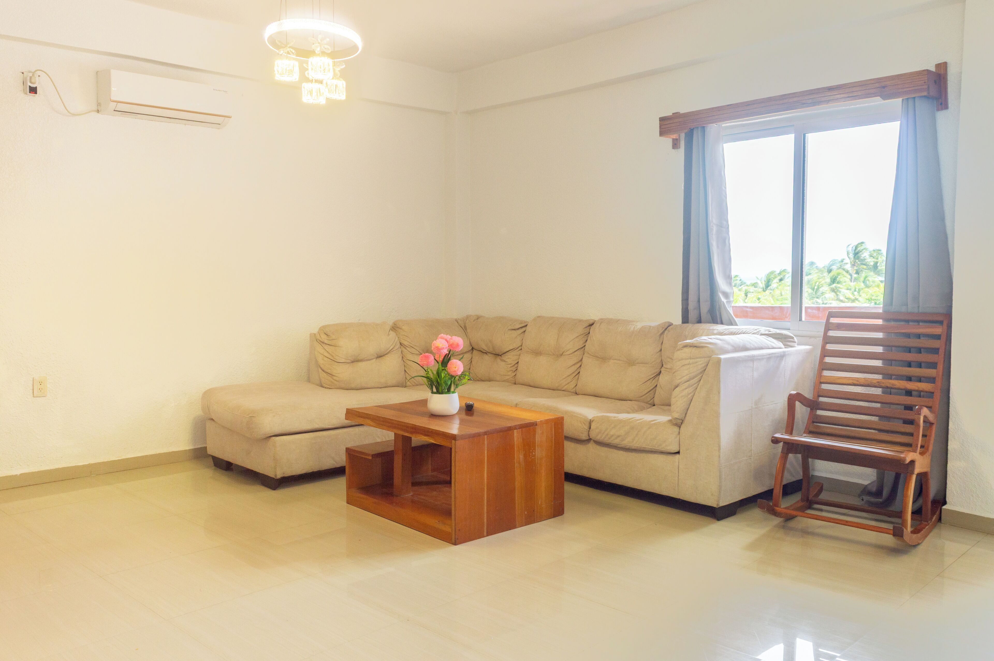 Una sala de estar con un sofá, una mesa de centro de madera con un jarrón de flores, un sillón mecedor de madera y un ventanal con cortinas.