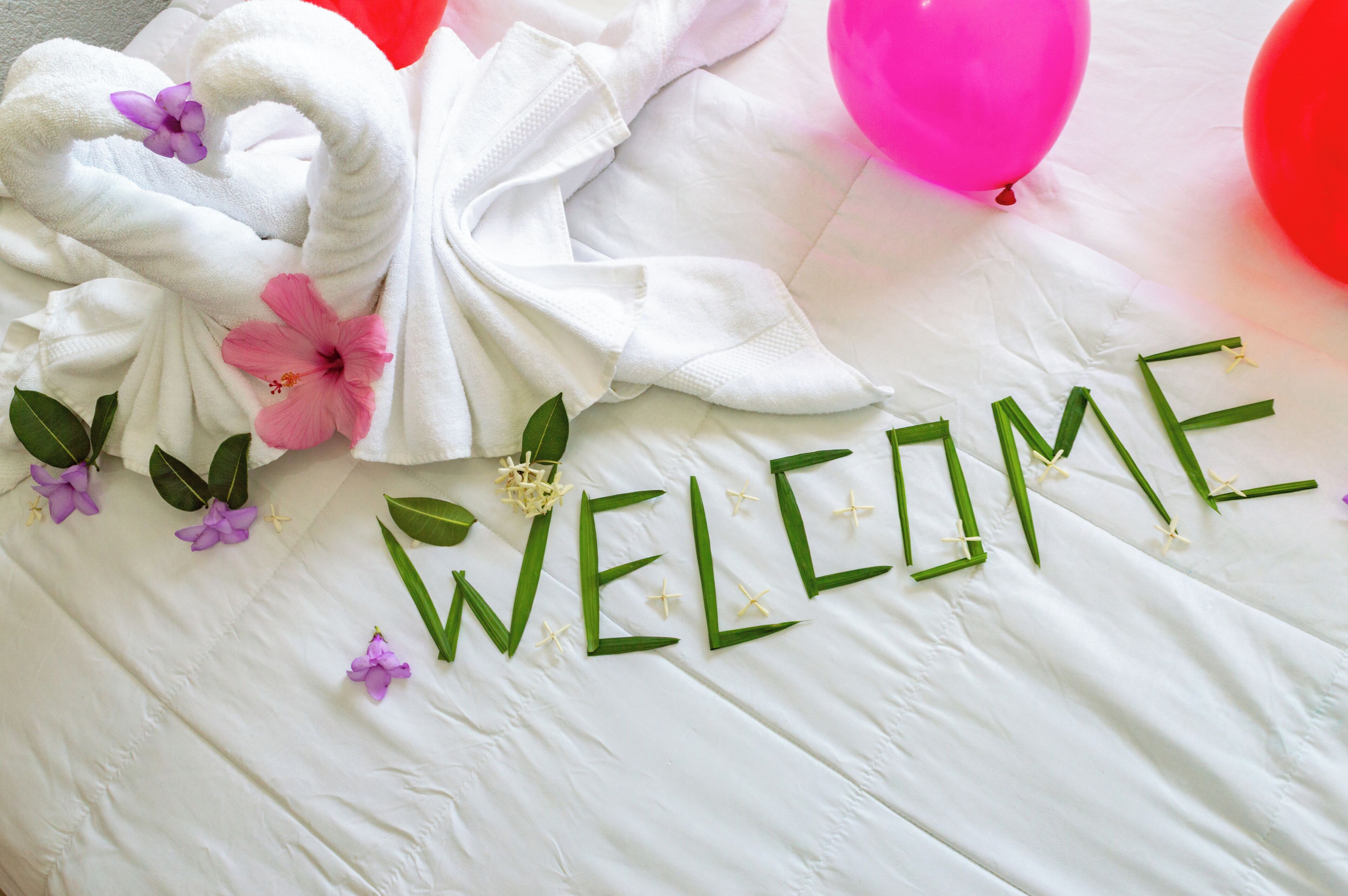 A white towel arranged in a swan shape with pink and purple flowers, and the word "WELCOME" spelled out with green leaves and flowers.