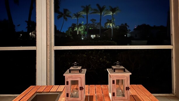 Una mesa de madera con dos faroles frente a una ventana que da a palmeras de noche.