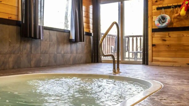 A wooden cabin interior with a hot tub, wooden walls, and a view of a wooden deck outside. - 3 Bedroom, Sleeps 10, Close to town w Pool, Hot Tub, Sauna and Game Room! (Sevierville)