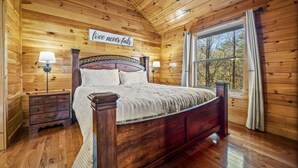 A wooden cabin bedroom with a bed, nightstands, and a window with curtains. - 3 Bedroom, Sleeps 10, Close to town w Pool, Hot Tub, Sauna and Game Room! (Sevierville)