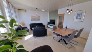 Uma sala de estar moderna com um sofá preto, uma mesa de jantar de madeira com cadeiras, uma TV e uma janela grande com cortinas.