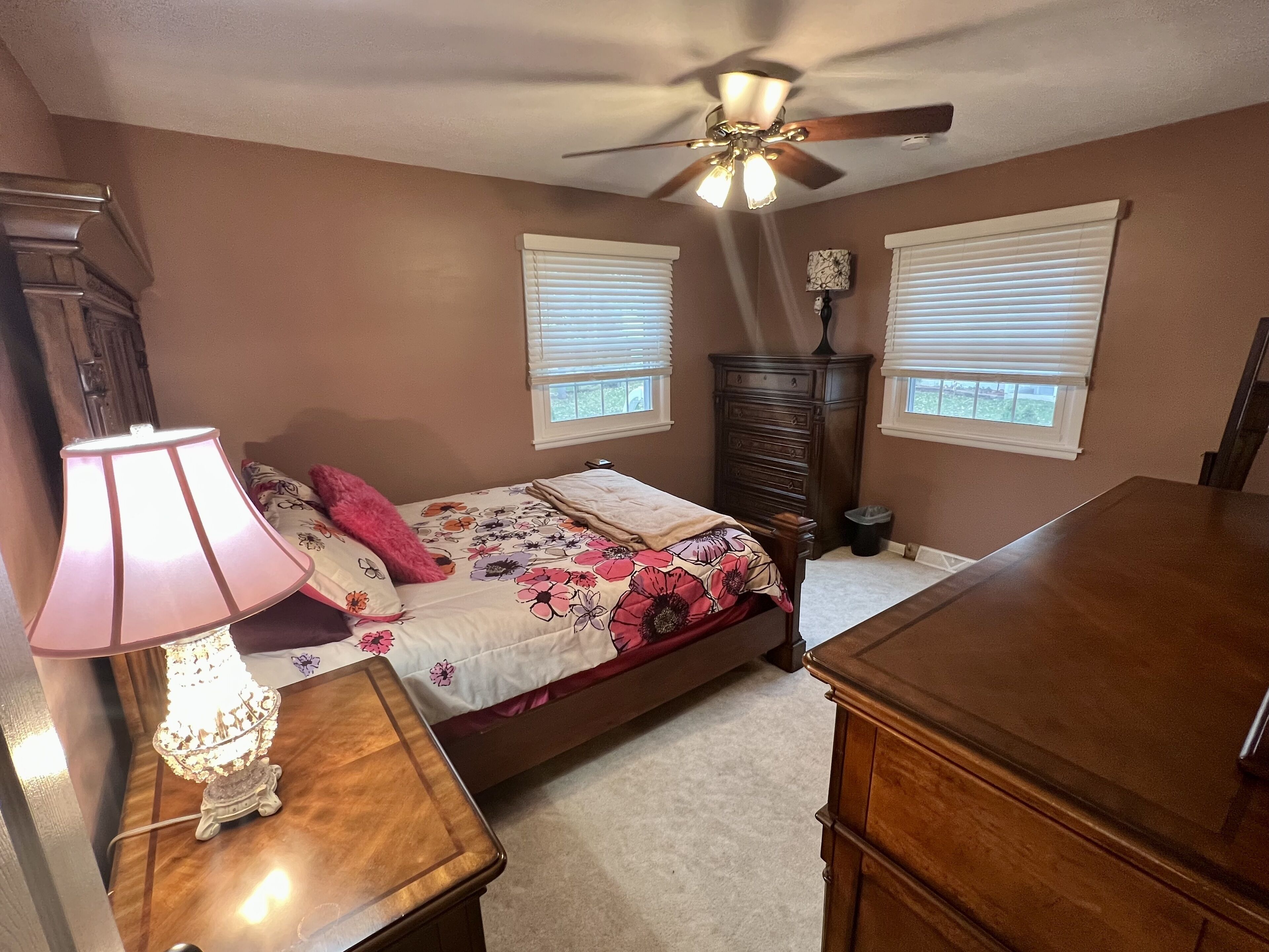 Une chambre à coucher comprenant un lit, une commode, une table de chevet, une lampe, un ventilateur de plafond et des fenêtres.