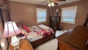 Une chambre à coucher comprenant un lit, une commode, une table de chevet, une lampe, un ventilateur de plafond et des fenêtres.