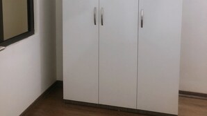 A white wardrobe with two doors and handles, positioned in a corner with a wooden floor and a white wall. - Selene Home (Hanoi)