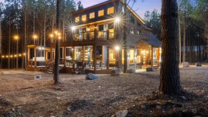 A two-story wooden cabin with a balcony, surrounded by trees and a gravel ground. - Fore Sight Golf Simulator, Games, Play-Set, Pets, Tee It Up (Broken Bow)