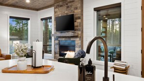 A modern kitchen with a stone fireplace, a flat-screen TV, and a large window overlooking a forest. - Fore Sight Golf Simulator, Games, Play-Set, Pets, Tee It Up (Broken Bow)