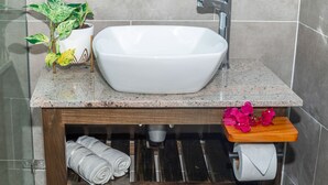 Un baño con un lavabo blanco tipo recipiente, una planta en maceta y un estante de madera con toallas y flores.