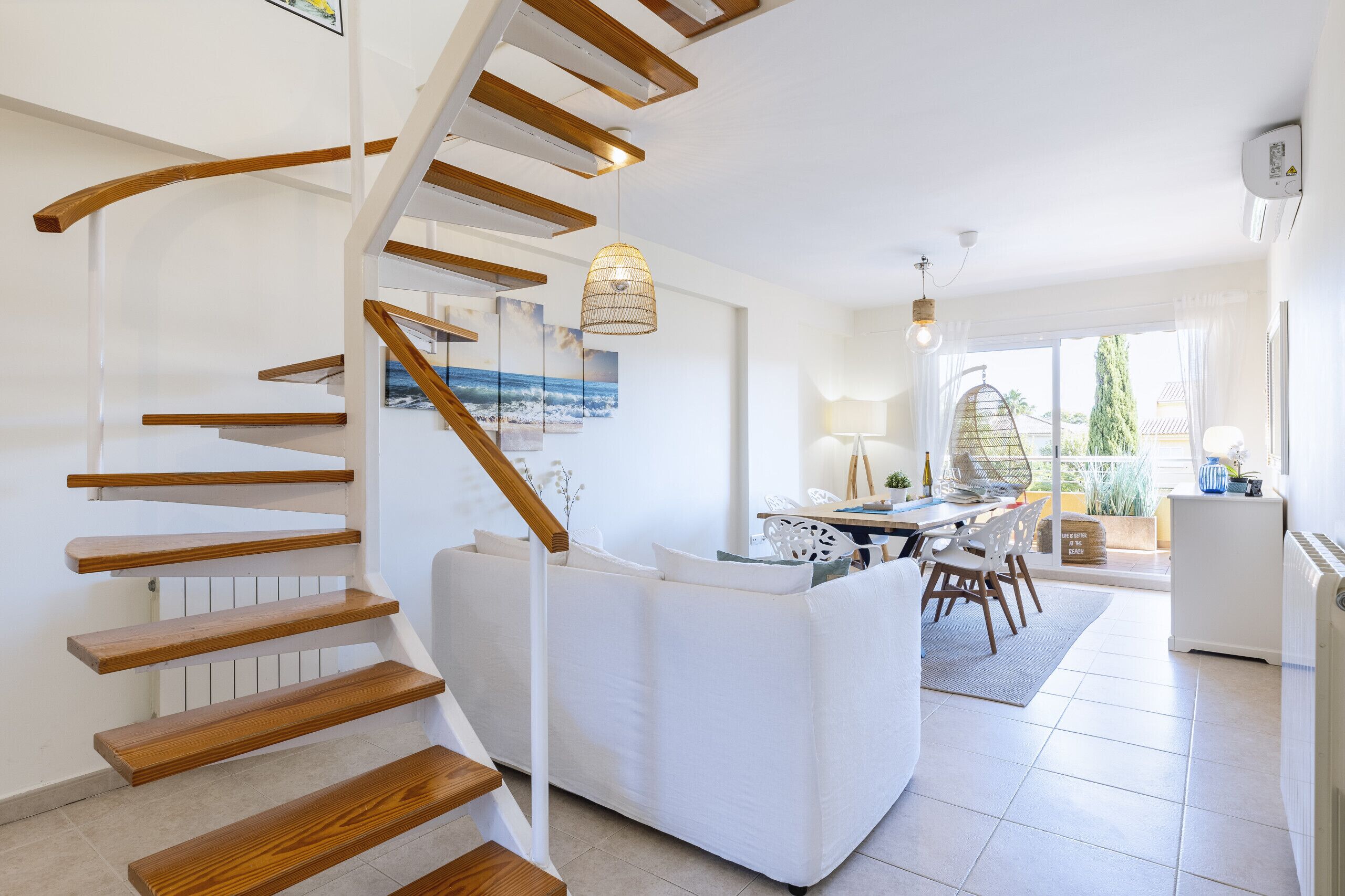 A modern interior with a wooden staircase, a white sofa, and a dining area with a view of the outdoors.