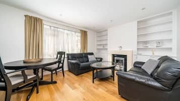 A modern living room with a black leather sofa, a glass coffee table, and a dining table with chairs. There is a fireplace and built-in shelving against the wall.