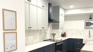 A modern kitchen with dark blue cabinets, a white countertop, and a stainless steel oven. There are framed pictures on the wall and a wooden dining table.