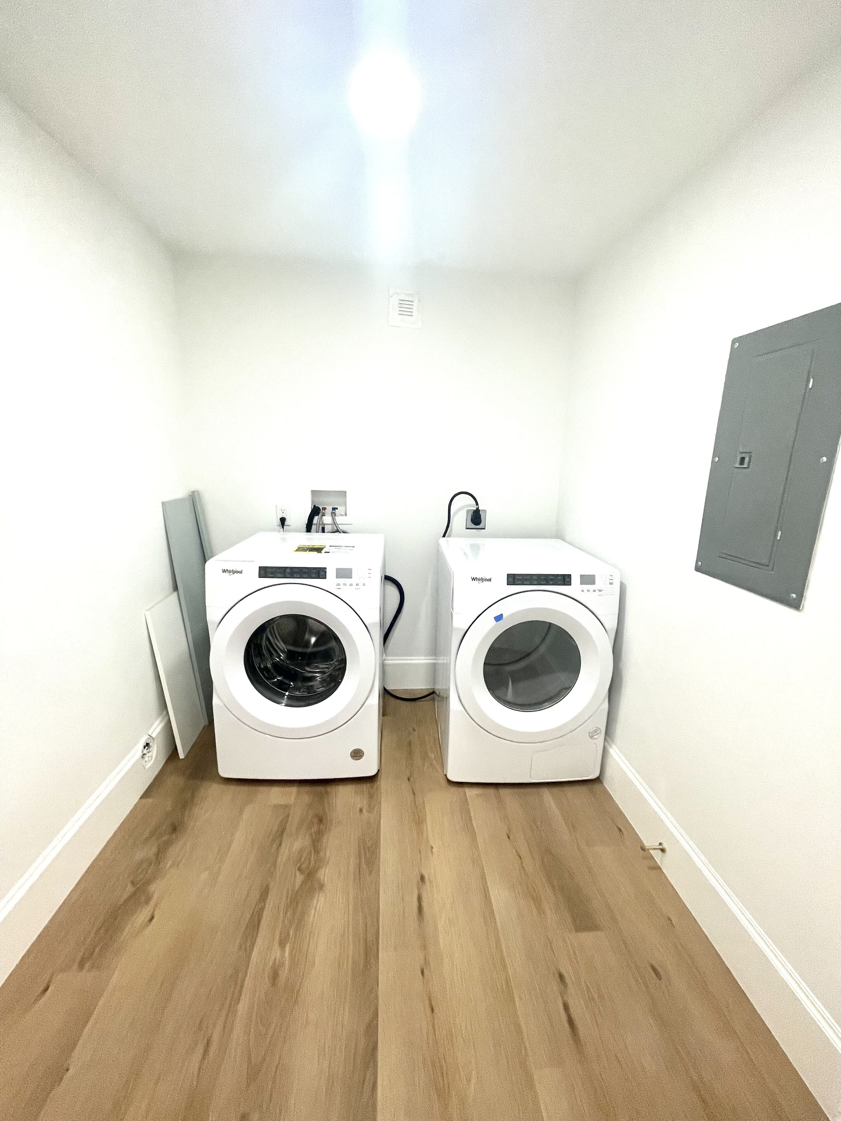 A laundry room with two white Whirlpool washers, a wooden floor, and a white wall with a small electrical panel.