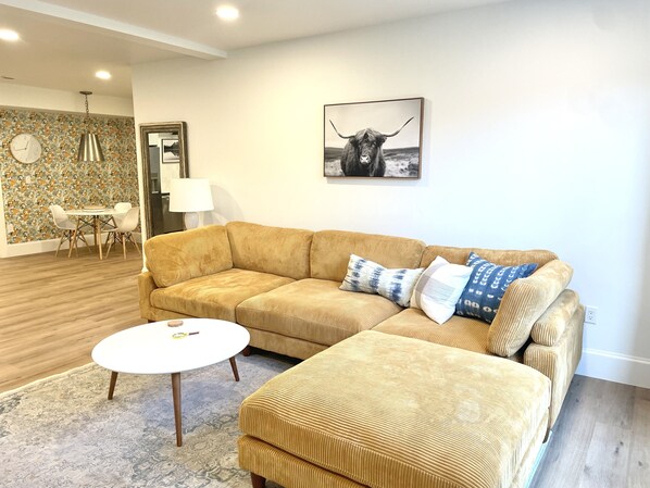 A living room with a large beige sectional sofa, a small white coffee table, and a dining area with a floral wallpaper and wooden table.