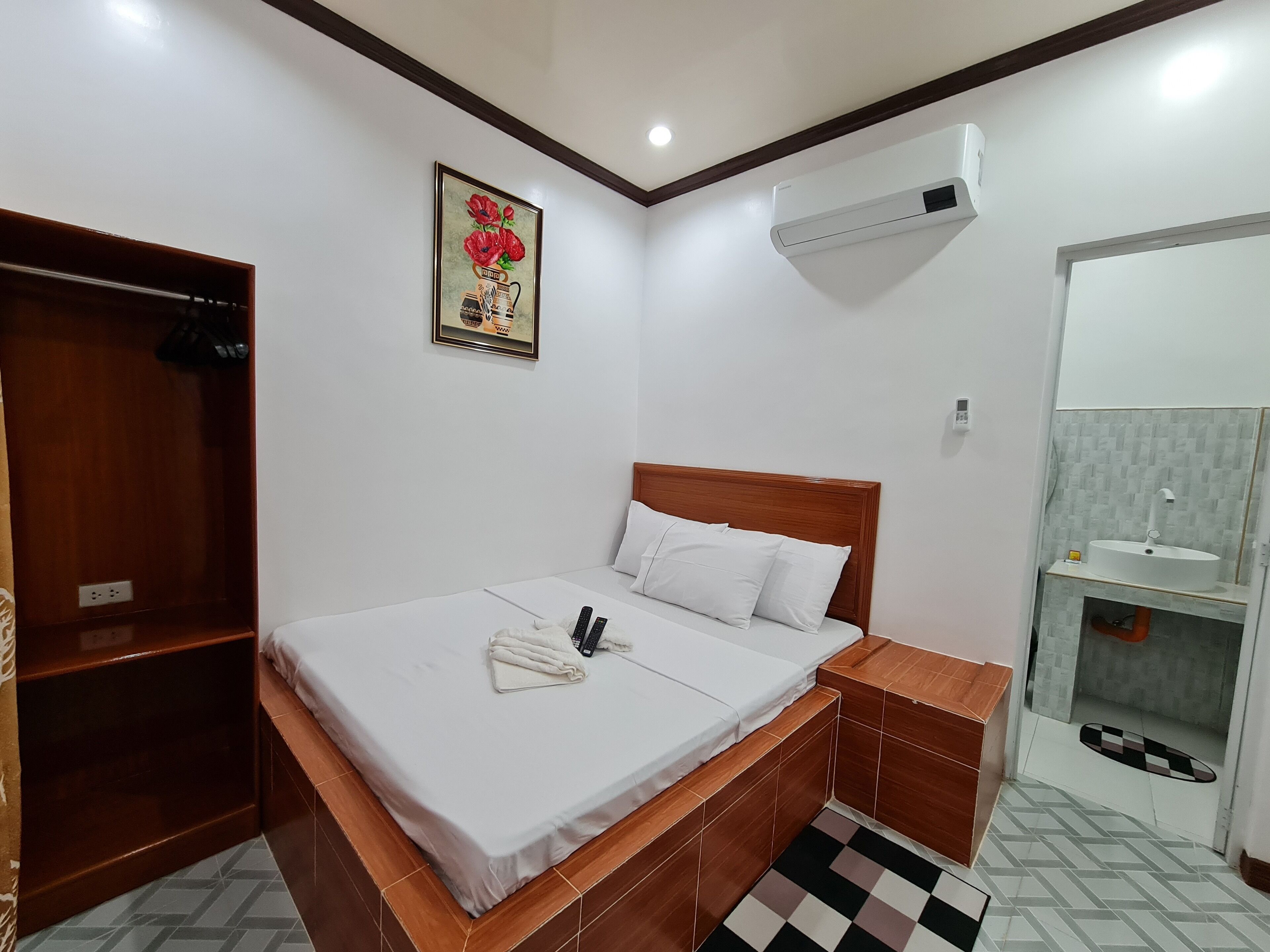 Un dormitorio con cama de madera, ropa de cama blanca, armario de madera, aire acondicionado instalado en la pared y un baño pequeño con lavabo y espejo.