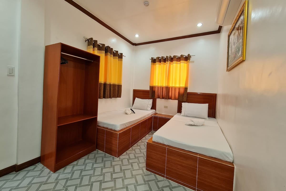 Une chambre d’hôtel avec deux lits simples, une armoire en bois et une fenêtre avec des rideaux jaunes.
