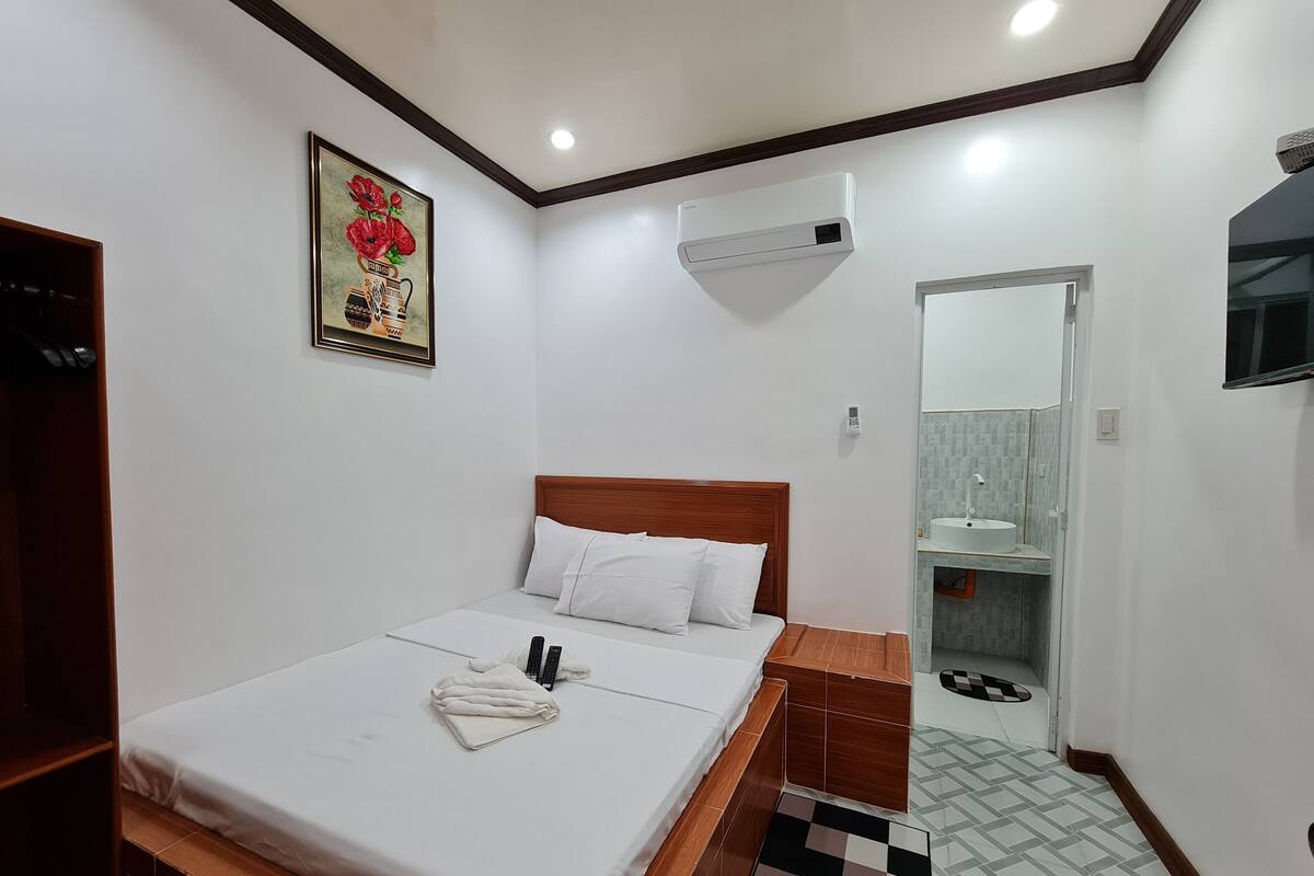 Une chambre d’hôtel avec un lit, une tête de lit en bois, un mur blanc, un appareil de climatisation, une salle de bain avec un lavabo et un miroir, et un sol carrelé.