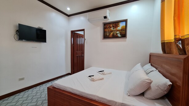 Un dormitorio con cama de madera, ropa de cama blanca, un televisor montado, un cuadro y una ventana con cortinas.