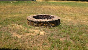 A stone fire pit in a grassy area with a building in the background. - Cozy cabin nestled in the Ozarks with beautiful views from the front porch. (Huntsville)