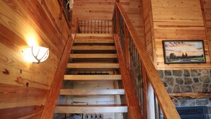 A wooden staircase with a metal railing, a stone fireplace, and a wall-mounted picture. - Cozy cabin nestled in the Ozarks with beautiful views from the front porch. (Huntsville)