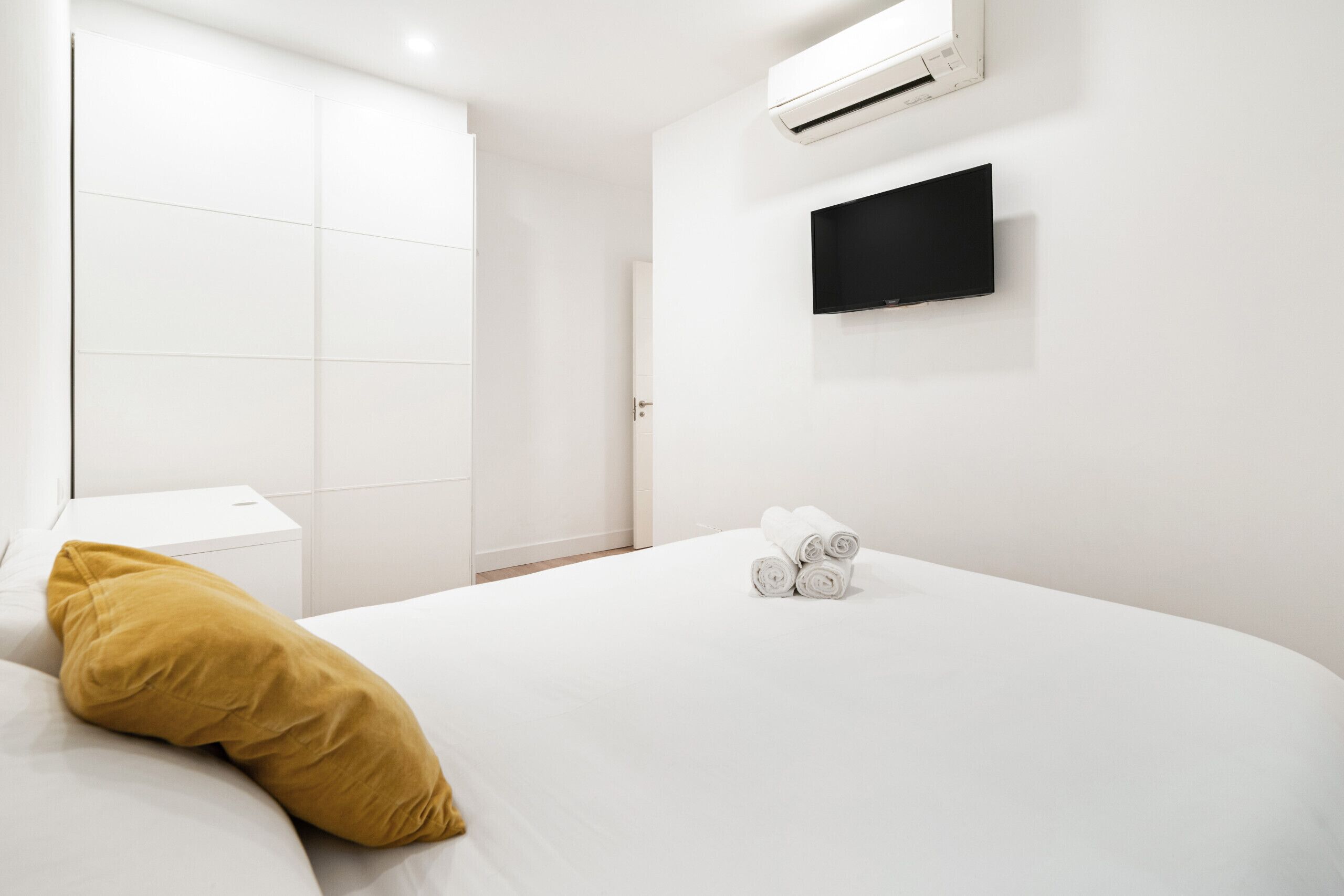 A hotel room with a bed, a white bedside table, a yellow pillow, a rolled-up towel, a flat-screen TV mounted on the wall, and an air conditioning unit.