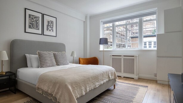 A bedroom with a large bed, a nightstand, a lamp, a window with a view of buildings, and a radiator. - Modern Bloomsbury Studio, nr Universities & Euston Station, by Blueground (London)