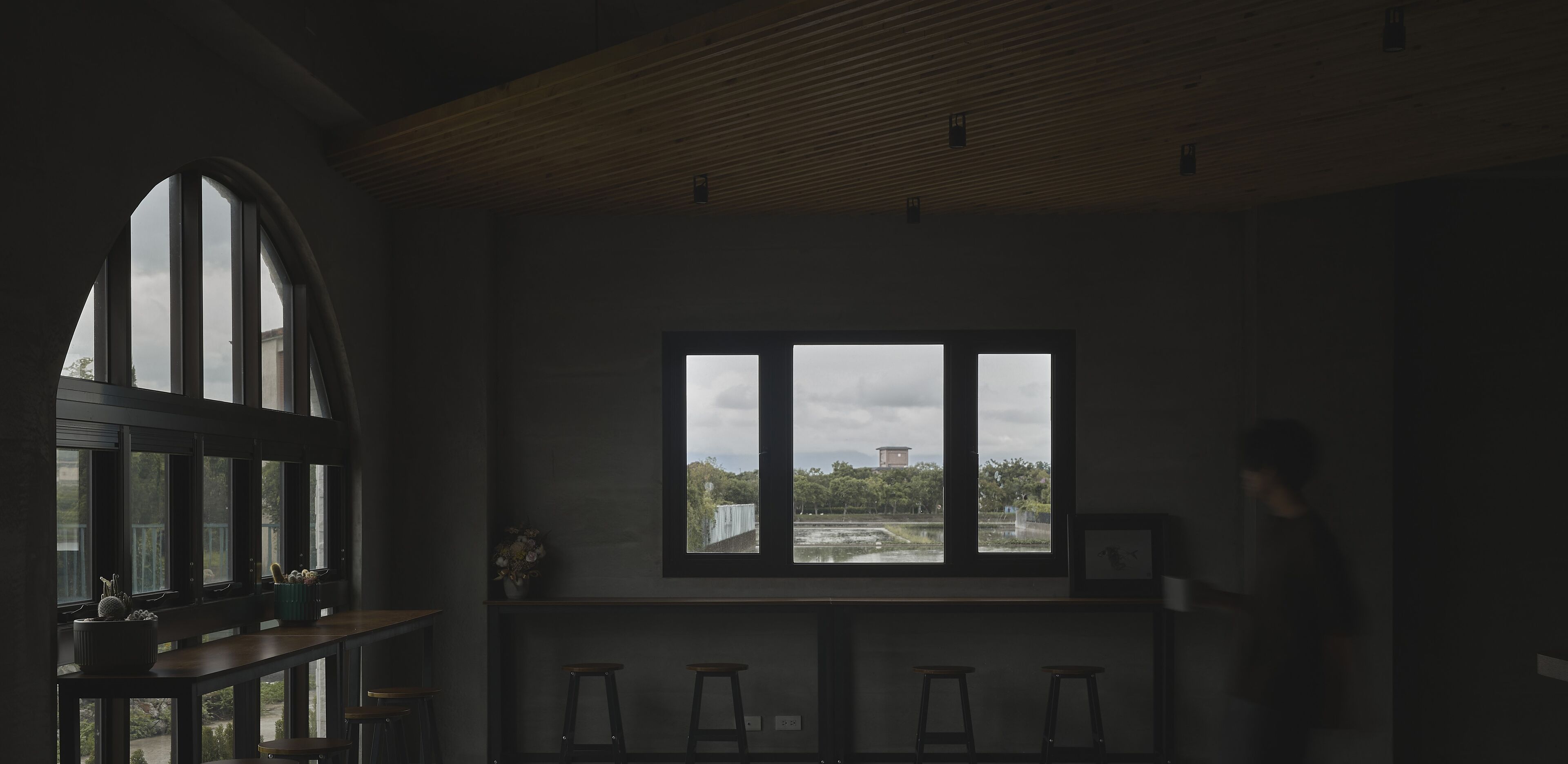 Een zwak verlichte kamer met een groot rondboogvenster, een houten plafond en uitzicht op een bewolkte lucht en verre gebouwen.