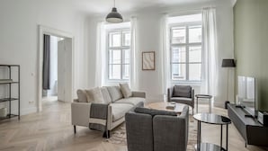 A modern living room with a sofa, armchairs, a coffee table, and a TV. There are large windows with curtains, a hanging light fixture, and a bookshelf.