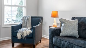 Ein Wohnzimmer mit einem dunkelblauen Sessel, einem grauen Sofa, einer Lampe und einem Fenster mit Jalousien.