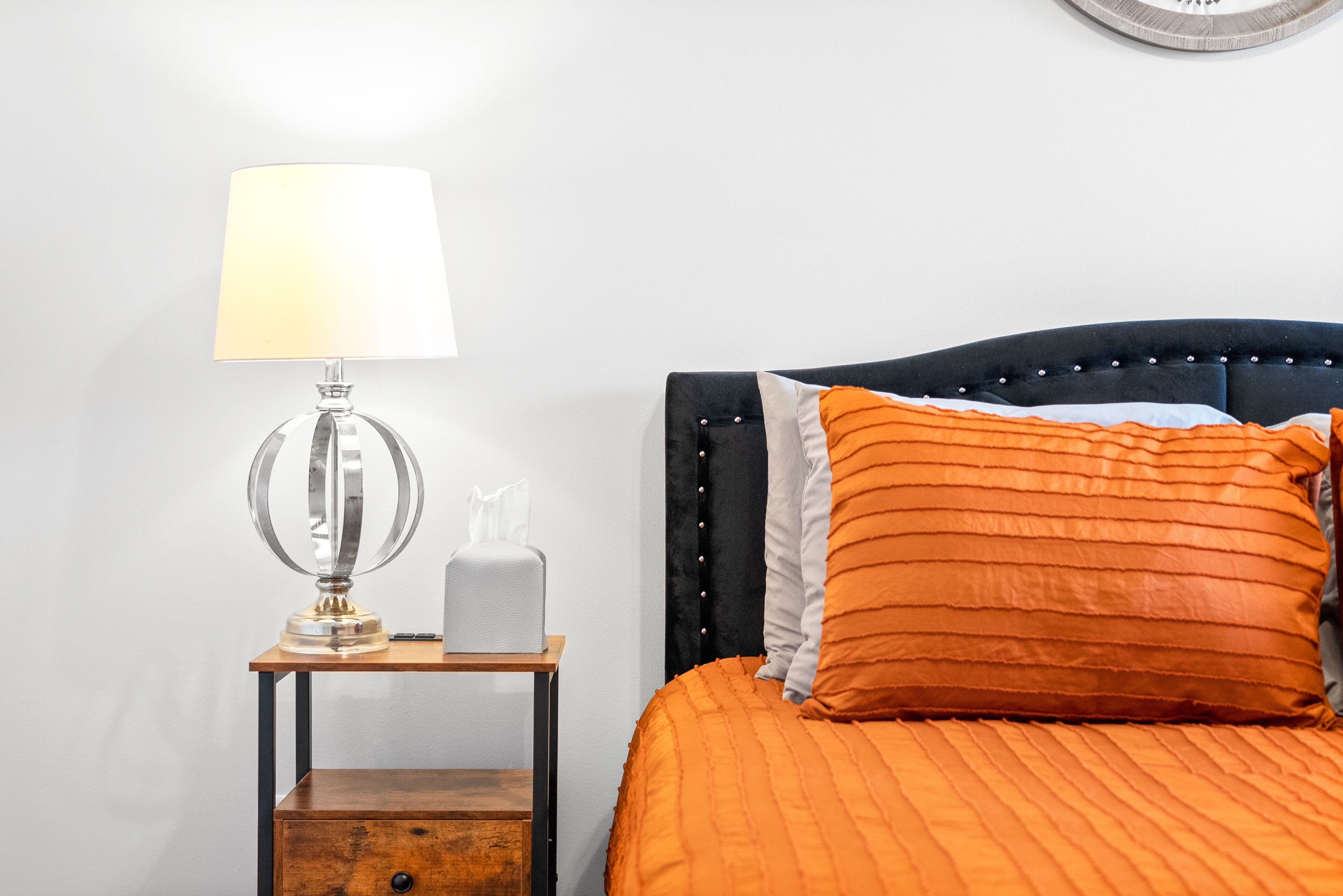 Uma cama bem arrumada com colcha laranja, uma luminária de cabeceira e uma pequena mesa de cabeceira de madeira com uma caixa de lenços.