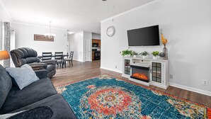 Ein Wohnzimmer mit einem dunkelgrauen Sofa, einem farbenfrohen Teppich, einem Kamin und einem Flachbildfernseher auf einer weißen Konsole.