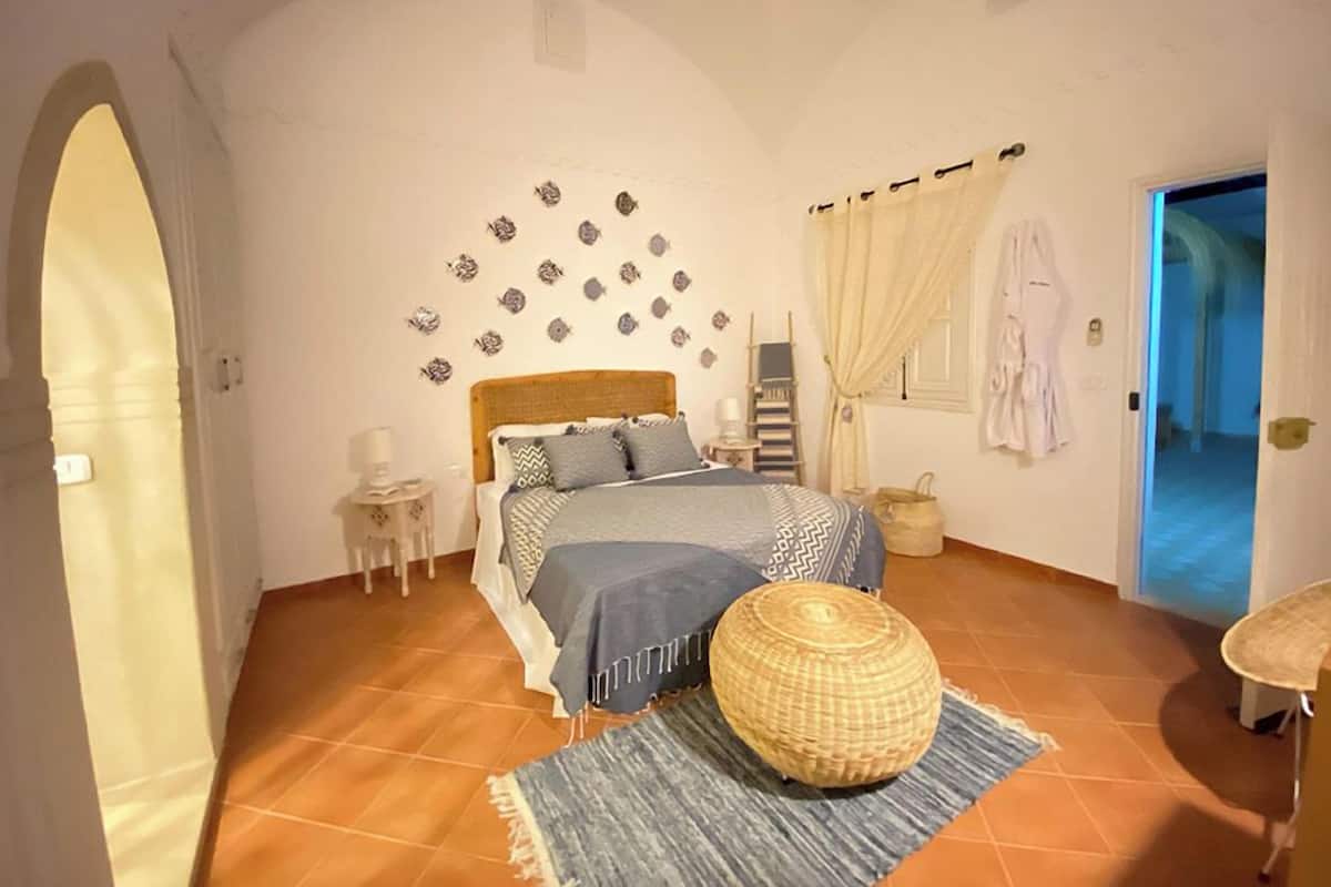 A bedroom with a bed, a small side table, a wicker ottoman, a blue rug, a wall-mounted shelf, and a door leading to another room.