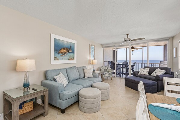 A living room with a sectional sofa, ottomans, a coffee table, and a lamp. There is a painting of a sea turtle on the wall. - Bay & Gulf Views | 5th Floor Condo, Pool & Walk to Times Square (Fort Myers Beach)