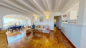 Uma sala ampla com sofá, mesa de jantar e cadeiras, cozinha com armários brancos e uma grande janela com vista.
