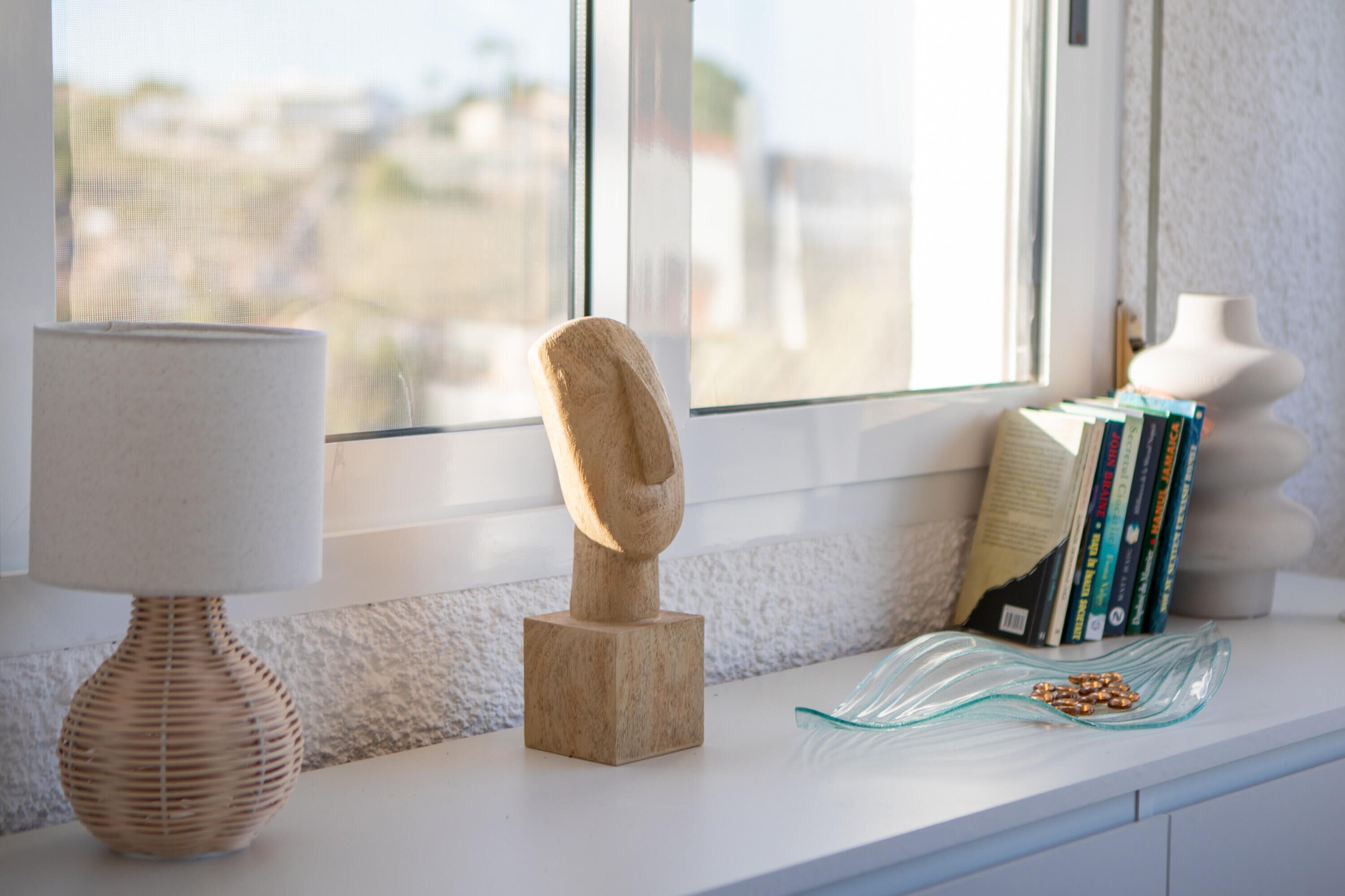 A room with a window, a lamp, a wooden sculpture, books, and a glass dish on a white surface.