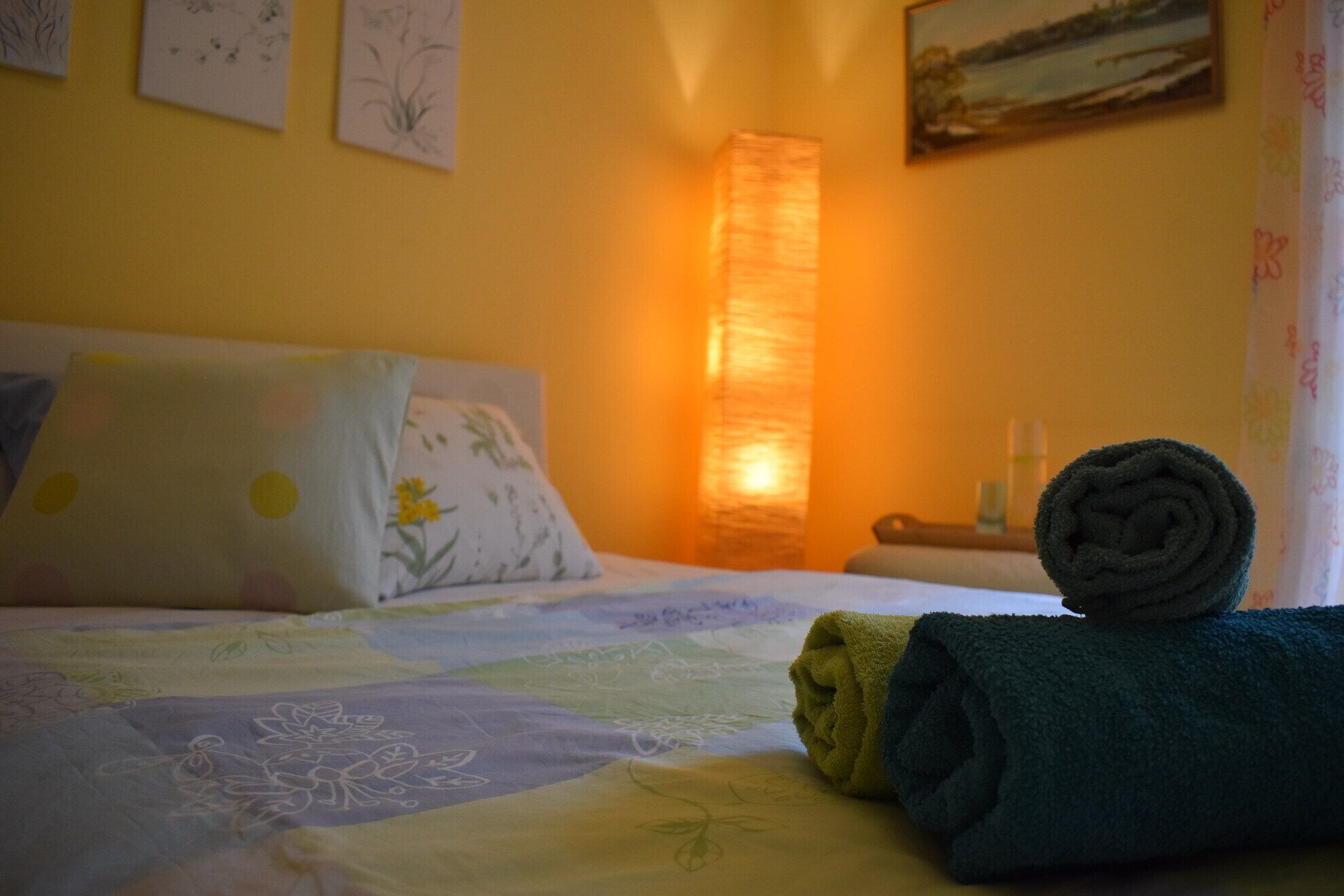 A bed with a floral-patterned bedspread, a rolled towel, and a nightstand with a bottle.