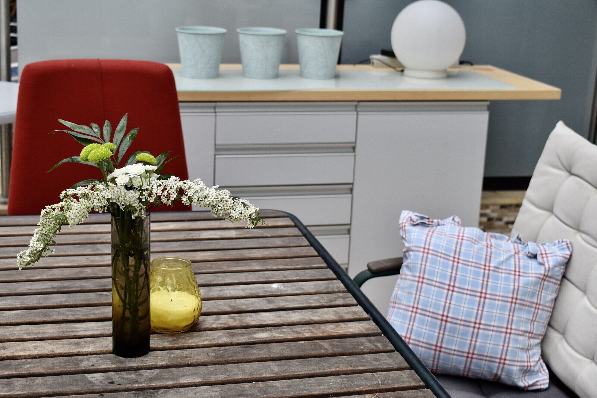 Une table en bois sur laquelle se trouve un vase de fleurs, un verre contenant un liquide jaune et un coussin à carreaux posé sur un banc.