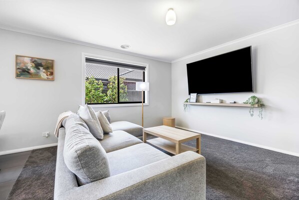 A modern living room with a sofa, a coffee table, a TV mounted on the wall, and a window with a view of greenery outside. - Spacious Living, Lucas (Lucas)