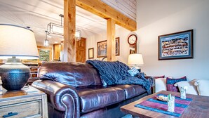A cozy living room with a leather sofa, wooden beams, a dining table with a placemat, and a large painting on the wall. - Elkhorn Ridge Condo + FREE Resort Amenities (Sun Valley)