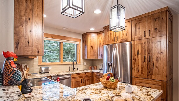 A kitchen with wooden cabinets, a stainless steel refrigerator, a granite countertop, and a window with a view of trees. - Elkhorn Ridge Condo + FREE Resort Amenities (Sun Valley)