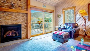 A cozy room with a fireplace, a leather sofa, a dining table with chairs, and a large sliding glass door leading to an outdoor patio. - Elkhorn Ridge Condo + FREE Resort Amenities (Sun Valley)