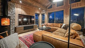 A cozy living room with a large flat-screen TV, a stone wall, wooden paneling, a leather sofa, and a red patterned rug. - Awesome home in Hornindal with WiFi (Hornindal)