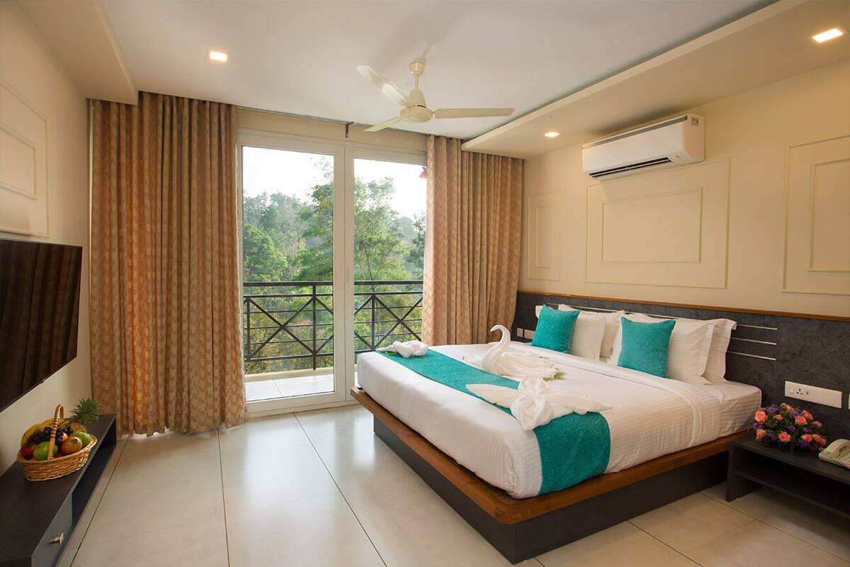Habitación de hotel con una cama grande, ventilador de techo, aire acondicionado, televisor y balcón con vistas a árboles.