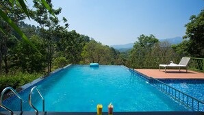 Une piscine avec vue sur une forêt et des montagnes, entourée d’une terrasse avec un fauteuil de relaxation et un flotteur.