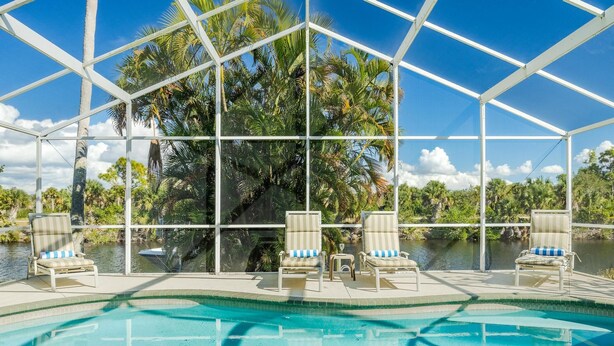 A pool area with clear blue water, surrounded by a glass enclosure with a view of palm trees and a body of water. - Newly Renovated, Gulf Access & Heated Pool - Villa Following the Sun - Roelens Vacations (Cape Coral)