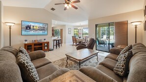 A spacious living room with a ceiling fan, a flat-screen TV, a dining area, and a sliding glass door leading to a patio. - Newly Renovated, Gulf Access & Heated Pool - Villa Following the Sun - Roelens Vacations (Cape Coral)